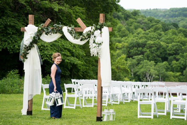 Blond Caucasian female wearing a formal navy bridesmaid dress and  holding two white lanterns walks under a draped canopy decorated for an outdoor wedding on a summer day, Minnesota, USA